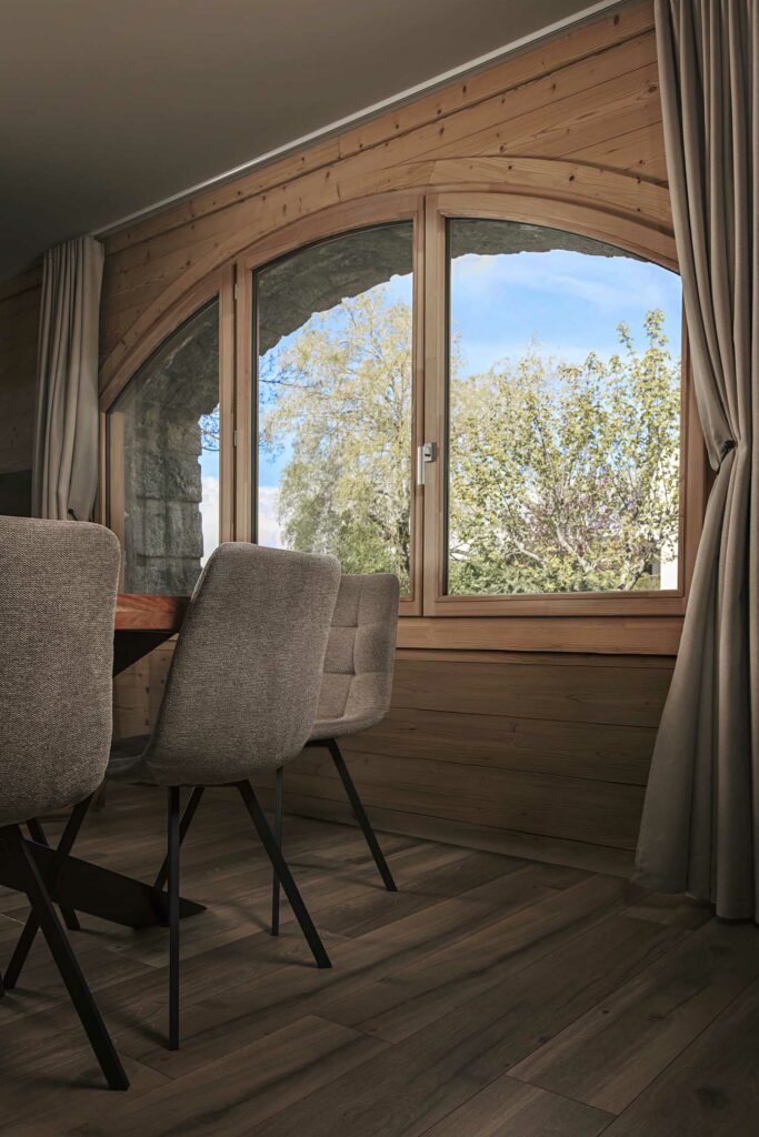 Salon chalet fenêtres cintrées bois, vue montagnes, canapé gris, plafond poutres apparentes