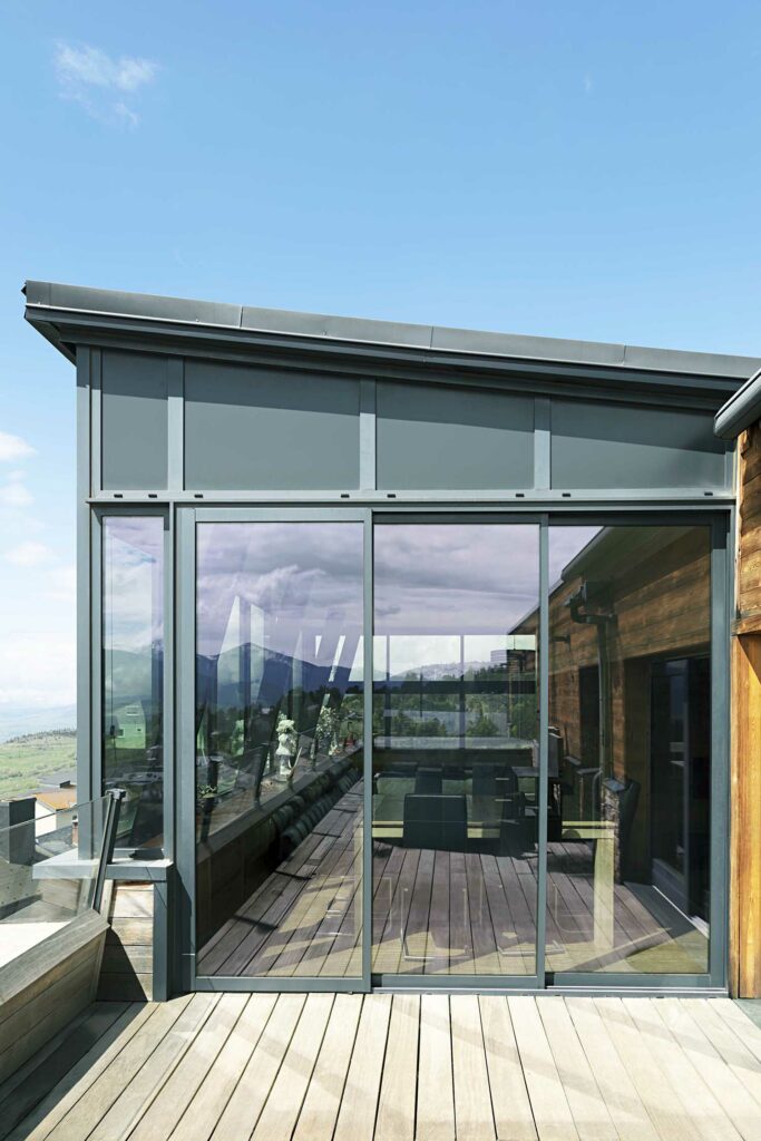 Véranda moderne avec structure aluminium gris et grandes baies vitrées, terrasse bois, vue paysage montagneux