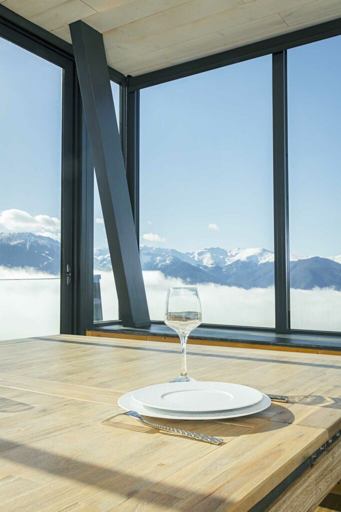Intérieur véranda haut de gamme avec baies vitrées panoramiques, table repas bois avec un verre de vin blanc et une assiette, vue montagnes enneigées, poutres bois naturel
