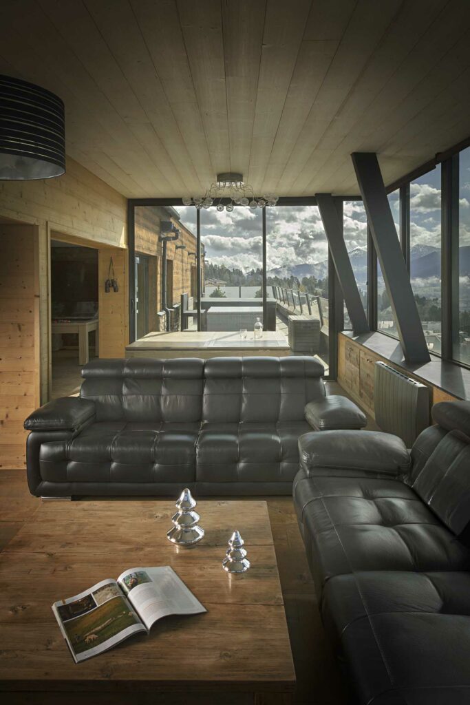 Salon véranda luxe avec canapé cuir noir, poutres inclinées, vue panoramique montagnes, luminosité naturelle maximale